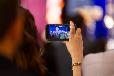 A woman holds a smartphone horizontally with one hand while recording a video at a graduation ceremony.